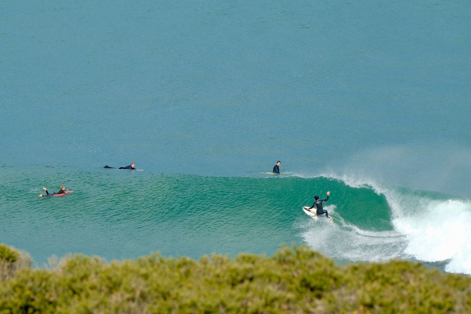 Surfer setting a rail on a breaking green wave as others paddle nearby